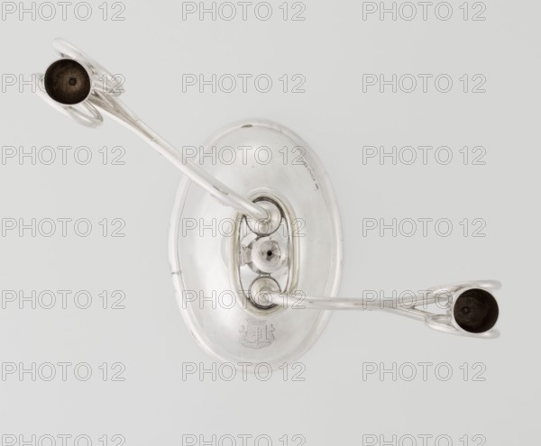 Two-candle candlestick, maker: William Leuchars fl. 1876–1888; firm: Leuchars & Son London; goldsmith's firm; fl. ca. 1845–1902. Silver candlestick with two movable arms made of S-shaped twisted wires, split at the ends, terminating in cylindrical candle sockets. Two-branched candlestick on a circular domed foot; shaft rests on a ball no finial and is enclosed at three-quarters of its height in a plate-like ring from which the arms extend. Foot engraved with the Pilawa coat of arms beneath a nine-baton count's crown and motto. Date: 1881–1882. Place of origin: London, England, Great Britain. Function: lighting. Materials/technique: goldsmithing/silver. Dimensions: height 27 cm; width 32 cm; depth 11.5 cm; weight 1082 g., A metallic wall-mounted fixture with an oval backplate and two curved arms extending outward, each arm ending in a shallow cup; the object is shiny silver with some darker interior areas in the cups and slight surface blemishes, set against a pale gray background.