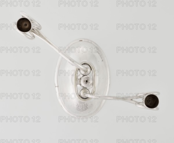 Two-candle candlestick, William Leuchars fl. 1876–1888, Leuchars & Son London; goldsmith's firm, fl. ca. 1845–1902. Two-branched candlestick on a circular domed foot; shaft rests on a ball no finial and is enclosed at 3/4 of its height in a plate-like ring, from which extend movable arms made of S-shaped twisted wires, split at the ends, terminating in cylindrical candle sockets. The foot is engraved with the Pilawa coat of arms with the motto beneath a nine-baton count's crown. Inscribed/marked: Pilawa coat of arms, Potocki coat of arms. Date: 1881/1882. Place of origin: London, England, Great Britain. Function: lighting. Materials/technique: goldsmith work. Dimensions: height 27 cm; width 32 cm; depth 11.5 cm; weight 1082 g., A top-down view of a metallic object with a smooth oval base and two curved arms extending outward, each ending in a small circular cup; the metal appears silver and reflective, with darker patina inside the cups, and the background is a pale light gray.