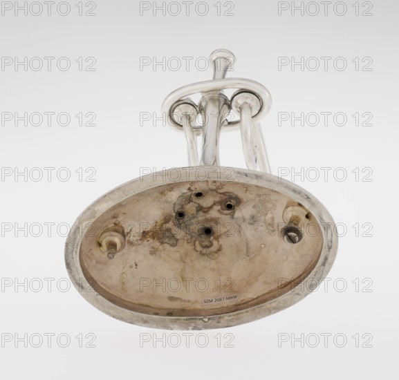 Two-candle candlestick by William Leuchars fl. 1876–1888, Leuchars & Son London; goldsmiths, fl. ca. 1845–1902. Two-branched candlestick on a circular domed foot; the shaft rests on a ball no finial, enclosed at 3/4 of its height in a plate-like ring, from which extend movable arms made of S-shaped twisted wires, split at the ends, terminating in cylindrical candle sockets. The foot is engraved with the Pilawa coat of arms with the motto beneath a nine-baton count's crown. 1881–1882. England, London. Lighting. Height 27 cm, width 32 cm, depth 11.5 cm, weight 1082 g., A metallic object viewed from below showing a curved oval base with a tarnished, stained surface and a few irregular dark spots and patches of discoloration; attached to the base are several shiny tubular metal rods and rounded fittings forming a looped structure above the base; visible colors include silver, gray, brown, beige, and darker rust-toned areas against a pale neutral background.