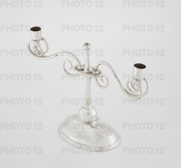 Two-candle candlestick. Maker: William Leuchars fl. 1876–1888; firm: Leuchars & Son London; goldsmiths; fl. ca. 1845–1902. Materials/technique: silver implied, lighting object. Description: two-branched candlestick on a circular domed foot; shaft rests on a ball no finial, enclosed at three-quarters of its height in a plate-like ring from which extend movable arms made of S-shaped twisted wires, split at the ends, terminating in cylindrical candle sockets. Foot engraved with the Pilawa coat of arms and motto beneath a nine-baton count’s crown; also bears Potocki coat of arms, rank crowns and mottos heraldry. Date: 1881–1882. Place of origin: London, England, Great Britain. Dimensions: height 27 cm, width 32 cm, depth 11.5 cm; weight 1082 g., A silver metal candelabrum with a rounded base and a tall central stem extends into two curved, scroll-like arms that each end in a cup-shaped candle holder; the metal appears polished and reflective, showing tones of silver and gray, with darker g...