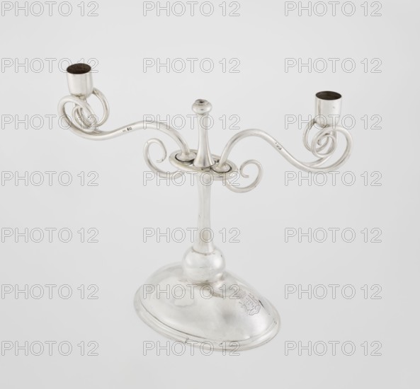 Two-candle candlestick by William Leuchars fl. 1876–1888, Leuchars & Son London; goldsmith's firm, fl. ca. 1845–1902. Two-branched candlestick on a circular domed foot; the shaft rests on a ball no finial and is enclosed at 3/4 of its height by a plate-like ring from which extend movable arms made of S-shaped twisted wires, split at the ends, terminating in cylindrical candle sockets. The foot is engraved with the Pilawa coat of arms with the motto beneath a nine-baton count's crown. 1881/1882. England, London. Lighting. Height 27 cm; width 32 cm; depth 11.5 cm; weight 1082 g., A silver-toned metal candelabrum with a rounded oval base and a tall central stem supporting a decorative horizontal ring and a group of curved, scroll-like arms ending in two cup-shaped candle holders, with visible reflective surfaces showing highlights and subtle tarnish in shades of silver and gray against a pale gray background.