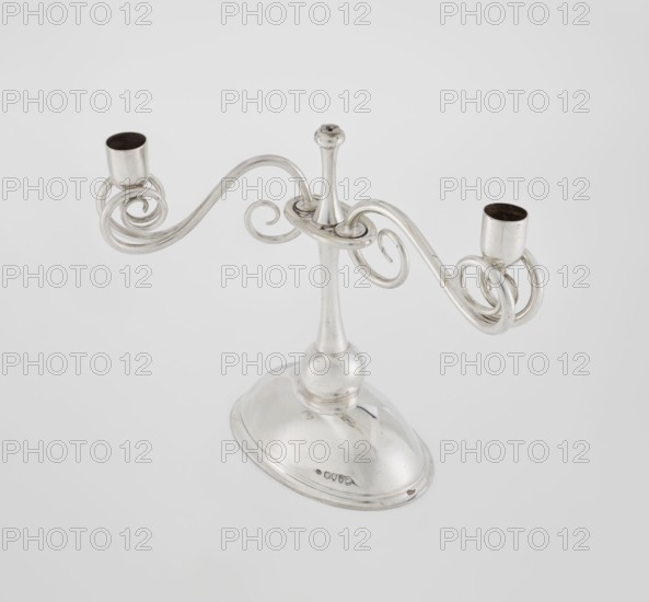 Two-candle candlestick. Maker: William Leuchars fl. 1876–1888, Leuchars & Son London; goldsmith's firm, fl. ca. 1845–1902. Materials/technique: silver implied, lighting object. Description: two-branched candlestick on a circular domed foot; shaft rests on a ball no finial, enclosed at three-quarters of its height in a plate-like ring from which extend movable arms made of S-shaped twisted wires, split at the ends, terminating in cylindrical sockets for candles. Foot engraved with the Pilawa coat of arms with motto beneath a nine-baton count's crown. Date: 1881–1882. Place of origin: London, England, Great Britain. Dimensions: height 27 cm; width 32 cm; depth 11.5 cm; weight 1082 g., A polished metal two-armed candelabrum with a rounded base and a central stem supporting two swirling curved arms ending in cylindrical candle holders; the metal appears silver with reflective highlights and darker interior surfaces in the holders, and the background is a pale light gray.
