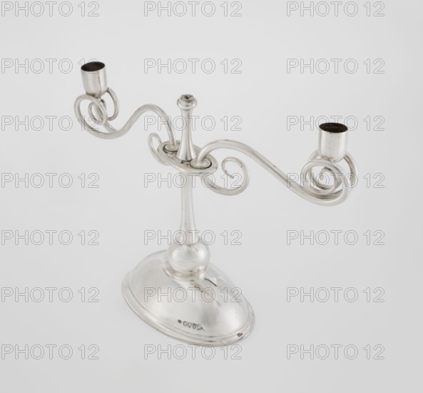 Two-candle candlestick by William Leuchars fl. 1876–1888 and Leuchars & Son London; goldsmith's firm, fl. ca. 1845–1902. Two-branched candlestick on a circular, domed foot. The shaft rests on a ball no finial, enclosed at three-quarters of its height in a plate-like ring, from which extend movable arms made of S-shaped twisted wires, split at the ends, terminating in cylindrical sockets for candles. The foot is engraved with the Pilawa coat of arms with the motto beneath a nine-baton count's crown. Materials/technique: scrap metal. Date: 1881/1882. Place of origin: London, England, Great Britain. Function: lighting. Dimensions: height 27 cm; width 32 cm; depth 11.5 cm; weight 1082 g., A silver-colored metal candelabrum with a rounded base and a tall, tapered central stem supporting a pair of ornate, curving arms that end in two cup-shaped holders; the arms form decorative spirals and loops and the metal surface appears reflective with some darker interior areas in the holders, set agai...