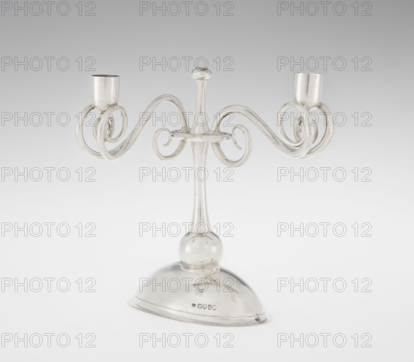 Two-candle candlestick, William Leuchars fl. 1876–1888, Leuchars & Son London; goldsmith's firm, fl. ca. 1845–1902. Two-branched candlestick on a circular domed foot. The shaft rests on a ball no finial, enclosed at three-quarters of its height by a plate-like ring from which extend movable arms made of S-shaped twisted wires, split at the ends, terminating in cylindrical candle sockets. The foot is engraved with the Pilawa coat of arms with the motto beneath a nine-baton count's crown. 1881–1882. England, London. Lighting object. Height 27 cm; width 32 cm; depth 11.5 cm; weight 1082 g., A clear glass candelabrum with a central stem rising from a rounded glass base, featuring a bulbous joint near the base and a slender neck topped by a small rounded finial, with two symmetrically curved arms that form ornate spiral loops ending in cylindrical candle cups; the colors visible are mainly transparent glass with reflections showing shades of white, pale gray, and subtle metallic highlights.