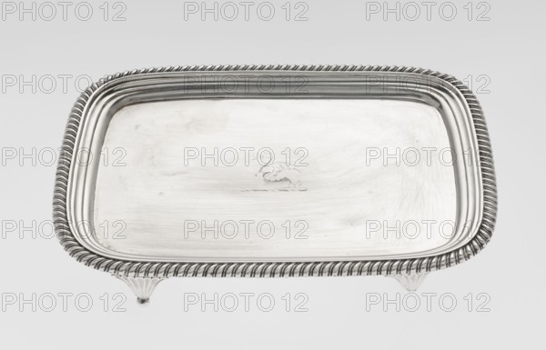 Tray on legs by William Bennet fl. 1796–1825, goldsmith. Tray on four legs in the form of stylised lotus flowers, rectangular with rounded edges, with a mirror, decorated with an engraved representation of a swan drawing heavily worn, with a raised, profiled collar and an edge decorated with a godron ribbon. Godron ornaments; English Regency style; swans iconography. Dated 1811/1812, London, Great Britain. Tray; height 3.3 cm, width 26.6 cm, depth 20.4 cm; weight 758 g., A rectangular metal tray with rounded corners sits on small decorative feet, the tray surface showing subtle brushed textures and a small engraved motif near the center; the tray rim features a twisted rope-like border and stepped inner edge, and the visible colors are silver, gray, and white.