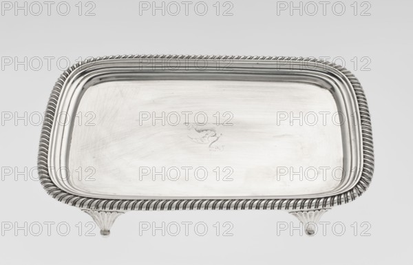 Tray on legs by William Bennet fl. 1796–1825, goldsmith. Tray on four legs in the form of stylised lotus flowers; rectangular with rounded edges and a mirror; decorated with an engraved representation of a swan drawing heavily worn; raised, profiled collar; edge decorated with a godron ribbon. Godron ornaments; English Regency style; swans iconography. Dated 1811/1812, London, Great Britain. Tray; height 3.3 cm, width 26.6 cm, depth 20.4 cm, weight 758 g., A rectangular metal tray with rounded corners sits on a plain light background, showing a shiny silver surface with a decorative twisted rope-like rim and a stepped inner border; the tray rests on a few short fluted feet and the surface displays faint scratches and subtle variations in silver tones.