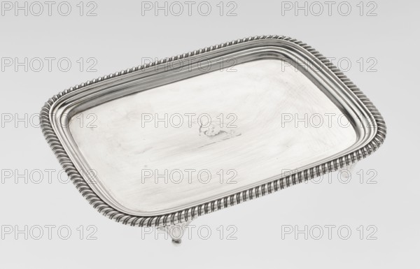 Tray on legs by William Bennet fl. 1796–1825, goldsmith. Tray on four legs in the form of stylised lotus flowers, rectangular with rounded edges, with a mirror, decorated with an engraved representation of a swan drawing heavily worn, with a raised profiled collar and an edge decorated with a godron ribbon. English Regency style, godron ornaments, swans iconography. Dated 1811/1812, London, England. Tray; height 3.3 cm, width 26.6 cm, depth 20.4 cm, weight 758 g., A rectangular metallic tray with rounded corners and a patterned raised rim sits on small ornate feet; the tray is primarily silver with reflective highlights and subtle darker gray areas from shadows and surface texture, and it shows a lightly brushed surface with faint scratches and a small decorative embossed motif near the center; the rim has a twisted rope-like detail and concentric raised bands, and the feet have fluted designs with rounded tips.