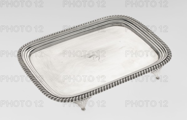 Tray on legs by William Bennet fl. 1796–1825, English Regency, 1811–1812. Rectangular tray with rounded edges on four legs shaped as stylised lotus flowers, with a mirror and raised profiled collar; edge decorated with a godron ribbon. Decorated with an engraved representation of a swan drawing heavily worn. Materials: presumably silver; height 3.3 cm, width 26.6 cm, depth 20.4 cm, weight 758 g., A rectangular metal tray with rounded corners sits on three short decorative feet; the tray surface and raised rim are metallic silver with a polished finish, showing fine scratches and subtle surface marks, and the rim features a repeating twisted rope-style pattern in silver.