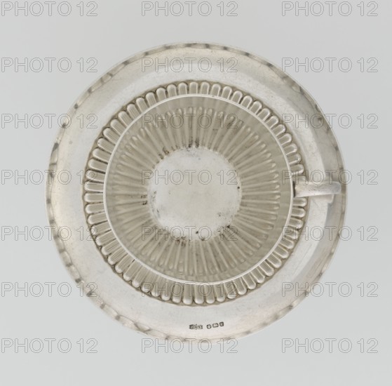 Hemispherical cup and saucer, decorated with radiating curls to half the cup's height, with a C-shaped side handle. Round saucer with a smooth rim and a mirror decorated with radiating curls. Maker: James Dixon & Sons Sheffield; goldsmiths; fl. 1806– . Date: 1891–1892. Place: Sheffield, England, Great Britain. Material/function: vessel, cup and saucer. Dimensions: cup height 6 cm, width 11 cm, depth 8.5 cm; saucer height 1.7 cm, diameter 14.2 cm. Weight: approx. 420 g., A round metallic plate with a matching shallow bowl centered on it, both showing a silvery metallic sheen with areas of darker tarnish and small spots of discoloration; the bowl has a ribbed radial pattern on its interior and a scalloped inner rim, and a small metallic handle extends from one side of the bowl; the background is a pale light gray, and the overall visible colors are silver, gray, and faint darker gray tones.