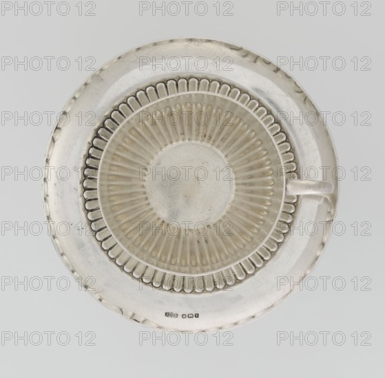 Hemispherical cup and saucer decorated with radiating curls to half its height; C-shaped side handle on cup. Round saucer with smooth rim and mirror decorated with radiating curls. Maker: Dixon, James & Sons Sheffield; goldsmiths; fl. 1806-. Date: 1891/1892. Place: Sheffield, England, Great Britain. Materials/role: goldsmiths. Function: vessel. Dimensions: cup height 6 cm, width 11 cm, depth 8.5 cm; saucer height 1.7 cm, diameter 14.2 cm. Weight approx. 420 g. Provenance: purchase., A top-down view of a circular metal cup and saucer set with a metallic silver color and slight tarnish; the saucer has a smooth rim with a few scuffs and a scalloped inner ring, and the cup shows a fluted pattern radiating from its center and a simple handle on the right side; the overall colors visible are silver, off-white, and faint gray.