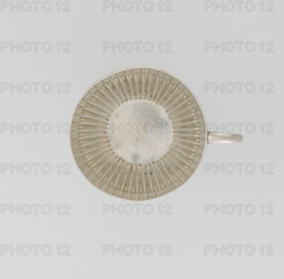 Hemispherical cup and saucer, decorated with radiating curls to half its height, with a C-shaped side handle. Round saucer with a smooth rim and a mirror decorated with radiating curls. Maker: Dixon, James & Sons Sheffield; goldsmiths; fl. 1806– . Date: 1891/1892. Place: Sheffield, England, Great Britain. Material/function: vessel, cup and saucer. Dimensions: cup height 6 cm, width 11 cm, depth 8.5 cm; saucer height 1.7 cm, diameter 14.2 cm. Weight approx. 420 g., A top-down view of a round metal cup with a ridged interior and a small handle extending to the right, showing colors including silver, gray, and off-white.