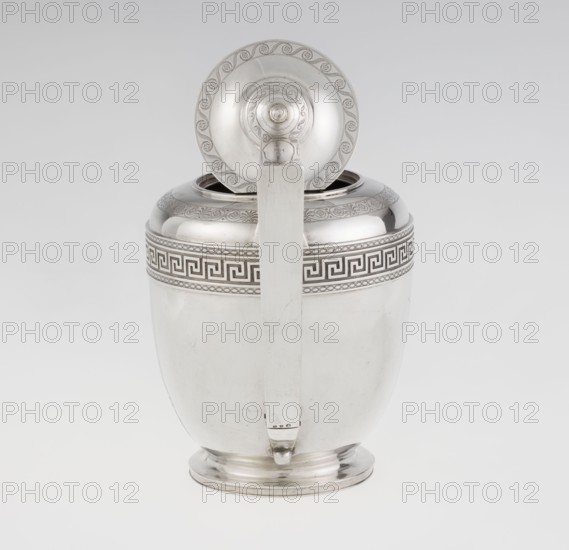 Teapot, maker: John Emes fl. 1796–1808. Ovoid silver teapot on a circular base with lid; Georgian/George III, Empire style, circa 1806–1807, London, England. Decorated on the upper belly with a wide band of meander ornament and braid; ribbon-shaped broken handle decorated with braid; high-set spout decorated at the base with a stylized palmette; lid finished with a knob-shaped handle. Belly engraved with a coat of arms on one side and a cross on the other. Vessel for tea. Height 17.9 cm, width 23.7 cm, weight 815 g., A polished metal container with a hinged lid shown open, featuring a vertical handle running down its front; the body is smooth and rounded with a slightly flared base and a decorative band near the top with a repeating angular pattern, and the lid and rim have circular ornamental engraving. The visible colors are silver, pale gray, and subtle reflections of white.