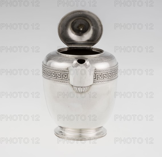 Teapot by John Emes fl. 1796–1808, silver, 1806–1807, London, England. Georgian/George III/Empire style. Ovoid teapot on a circular base with lid; upper belly decorated with a wide band of meander ornament and braid. Ribbon-shaped broken handle decorated with braid; high-set spout decorated at the base with a stylized palmette; lid finished with a knob-shaped finial. Belly engraved with a coat of arms on one side and a cross on the other. Tea vessel. Height 17.9 cm; width 23.7 cm; weight 815 g., A shiny metal vessel with a rounded body and narrow base sits against a plain light gray background, its hinged lid open to reveal a dark interior; the vessel is mostly silver in color with a decorative band near the top featuring a repeating angular pattern in darker silver, and a small protruding spout area with a scalloped detail beneath it, while the lid interior shows curved reflective surfaces and a central fastening element.