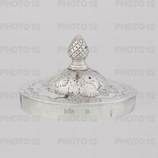 Buliera kettle for boiling water, maker: Benjamin Gignac fl. 1745–1774, goldsmith; rocaille ornament, rococo style, Georgian/George II influences; plant and floral ornament. Rococo kettle, spherical with a flat bow mounted on volutes; side spout; centrally arched cover topped with a pine cone; decorated with repoussé rococo decoration and flowers; includes two engraved coats of arms — one four-field in an oval under a baronial crown, the second in an illegible shield. Missing base with a heater. Dated 1753/1754, London, England, Great Britain. Vessel; height 22 cm, width 24 cm, depth 19 cm, weight 848 g., A metallic object with a reflective silver surface sits against a pale gray background; it has a rounded base with a low rim and a domed lid that rises to a textured finial shaped like a pinecone. The lid and dome show engraved floral and leafy patterns with areas of darker tarnish and subtle shadows that emphasize the embossed details. The overall colors visible are silver, gray, and...