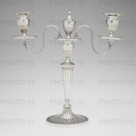 Two-candle candelabrum by Charles Hougham fl. 1769–1793. Two-armed candelabrum on an oval foot, bordered with pearling and decorated with a knobbed rosette. Conical, fluted stem topped with a vase-shaped socket, with a setting of two S-shaped arms terminating in saucers and vase-shaped sockets; in the centre between the arms is a vase topped with a ball. Sleeves decorated halfway with vertical flutes, smooth above. Classicism/George III/Georgian style; fluted, pearling, rosette ornament. Dated 1787/1788. England, London. Lighting. Height 38 cm; width 39 cm; depth 12 cm; weight 1060 g., A silver-colored three-armed candelabrum with a fluted round base and a tall, tapered fluted stem leading to a central urn-shaped holder topped by a removable lid, the central platform connecting to two curved, symmetrical arms that extend outward to hold a group of three cup-shaped candle sockets each set above a round drip plate; the metal surface is smooth and reflective with decorative fluting and be...