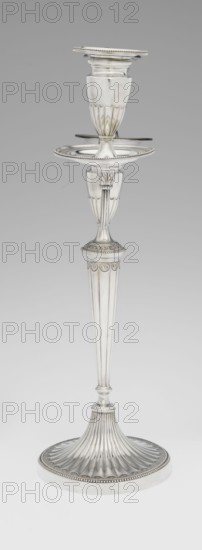 Two-candle candelabrum, Hougham, Charles fl. 1769–1793 goldsmith, classicism, George III, Georgian. Two-armed candelabrum on an oval foot, bordered with pearling and decorated with a knobbed rosette. Conical, fluted stem topped with a vase-shaped socket, in which is set a setting with two S-shaped arms terminating in saucers and vase-shaped sockets with drip pans; in the centre between the arms is a vase topped with a ball; sleeves decorated halfway with vertical flutes, smooth above. 1787/1788. England, London, Great Britain. Lighting. Height 38 cm; width 39 cm; depth 12 cm; weight 1060 g., A tall, slender silver candlestick with fluted and reeded details, showing a round, ridged base that fans upward into a tapered column with vertical grooves, a decorative collar with a band of repeating scallop-like motifs, a midsection featuring an ornate knop and a shallow circular dish with a beaded rim, and a cup-shaped holder at the top with fluted lower surface and a beaded edge; the visible ...
