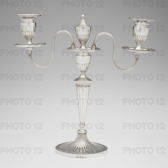 Two-candle candelabrum, Hougham, Charles fl. 1769–1793 goldsmith; classicism, George III/Georgian style; two-armed candelabrum on an oval foot bordered with pearling and decorated with a knobbed rosette. Conical, fluted stem topped with a vase-shaped socket, with a setting of two S-shaped arms terminating in saucers and vase-shaped sockets; centre between the arms a vase topped with a ball; sleeves decorated halfway with vertical flutes, smooth above. 1787/1788, London, England. Function: lighting. Height 38 cm, width 39 cm, depth 12 cm, weight 1060 g., A silver three-armed candelabrum stands against a neutral gray background, with a tall fluted central stem rising from a domed circular base that has radiating fluted grooves and a beaded rim; the central column features vertical fluting and decorative beaded and scalloped bands, leading to an urn-shaped central cup topped by a small domed finial. Two curved scrolling arms extend symmetrically from the central platform, each ending in a...