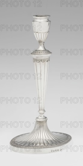 Two-candle candelabrum by Charles Hougham fl. 1769–1793, goldsmith. Classicism/Georgian, George III style. Two-armed candelabrum on an oval foot bordered with pearling and decorated with a knobbed rosette. Conical, fluted stem topped with a vase-shaped socket containing a setting with two S-shaped arms terminating in saucers and vase-shaped sockets; central vase topped with a ball. Sleeves decorated halfway with vertical flutes, smooth above. Dated 1787/1788. England, London. Lighting. Height 38 cm, width 39 cm, depth 12 cm, weight 1060 g., A tall metallic candlestick with a polished silver surface, featuring a rounded cup at the top with vertical fluted detailing below it, a narrow stem with ornamental rings and a decorative collar with scalloped motifs, a long tapered fluted column leading to a rounded joint, and a broad flared base with radiating flutes and a beaded rim; visible colors include silver and shades of gray against a light gray background.