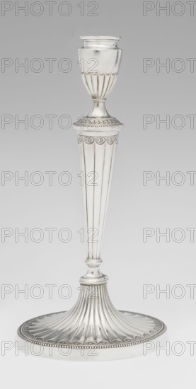 Two-candle candelabrum by Charles Hougham fl. 1769–1793. Classicism/George III Georgian style; fluted, pearling and rosette ornament. Two-armed candelabrum on an oval foot bordered with pearling and decorated with a knobbed rosette. Conical fluted stem topped with a vase-shaped socket holding a setting with two S-shaped arms terminating in saucers and vase-shaped sockets; central vase topped with a ball; sleeves decorated halfway with vertical flutes, smooth above. Dated 1787/1788. England, London. Function: lighting. Height 38 cm, width 39 cm, depth 12 cm, weight 1060 g., A tall metallic object with a silvery finish stands against a neutral gray background; it has a rounded cup-like top with vertical fluted details, a narrow stem with ornate circular and scalloped decoration, a long tapered column featuring vertical fluting, and a flared circular base with radiating ridges and a beaded edge, all showing reflective silver tones and subtle gray shadows.