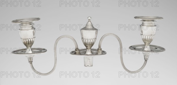 Two-candle candelabrum by Charles Hougham fl. 1769–1793, goldsmith, classicism/George III/Georgian style, 1787–1788, London, England. Two-armed candelabrum on an oval foot bordered with pearling and decorated with a knobbed rosette. Conical, fluted stem topped with a vase-shaped socket that holds a setting with two S-shaped arms terminating in saucers and vase-shaped sockets; central vase topped with a ball. Sleeves decorated halfway with vertical flutes, smooth above. Materials: metal silver; function: lighting. Dimensions: height 38 cm, width 39 cm, depth 12 cm; weight 1060 g., A silver metallic three-armed candleholder with a central covered cup and two matching side cups connected by curved arms, all with rounded bases and fluted lower sections; the surface is reflective with highlights and subtle shadows, showing tones of silver, gray, and off-white, set against a plain light gray background.