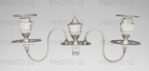 Two-candle candelabrum by Charles Hougham fl. 1769–1793, goldsmith; classicism/George III/Georgian style; 1787/1788, London, England. Two-armed candelabrum on an oval foot bordered with pearling and decorated with a knobbed rosette. Conical, fluted stem topped with a vase-shaped socket containing a setting with two S-shaped arms terminating in saucers and vase-shaped sockets with spouts; a central vase topped with a ball between the arms. Sleeves decorated halfway with vertical flutes, smooth above. Material: silver lighting. Dimensions: height 38 cm, width 39 cm, depth 12 cm; weight 1060 g., A symmetrical metallic object with a polished silver appearance rests against a neutral gray background, featuring a central covered urn with a rounded finial and fluted lower body, flanked by two curved double-armed supports that extend outward to hold matching open cup-like holders with fluted lower sections and flat circular drip trays with beaded edges; the metal shows reflective highlights an...