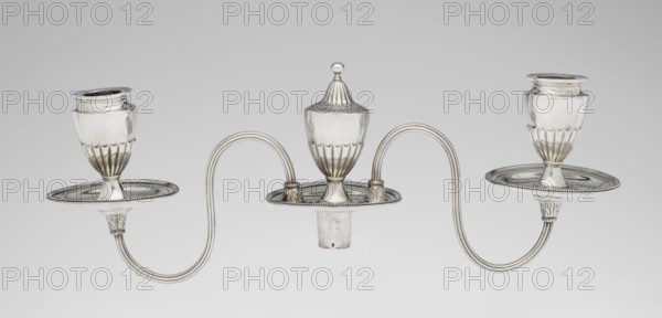 Two-candle candelabrum, Hougham, Charles fl. 1769–1793 goldsmith, classicism, George III / Georgian style. Two-armed candelabrum on an oval foot bordered with pearling and decorated with a knobbed rosette. Conical, fluted stem topped with a vase-shaped socket, containing a setting with two S-shaped arms terminating in saucers and vase-shaped sockets; in the centre between the arms is a vase topped with a ball. Sleeves decorated halfway with vertical flutes, smooth above. Scrap - purchased. 1787/1788. England, London. Function: lighting. Height 38 cm; width 39 cm; depth 12 cm; weight 1060 g., A silver three-armed ornamental object with an overall metallic sheen consisting of a central urn-shaped piece topped by a domed finial and two matching goblet-like holders on either side, connected by gracefully curved tubular arms that form symmetrical loops; each holder sits on a shallow round drip plate with beaded edges, and the bases of the supports feature fluted and acanthus-like decorative...