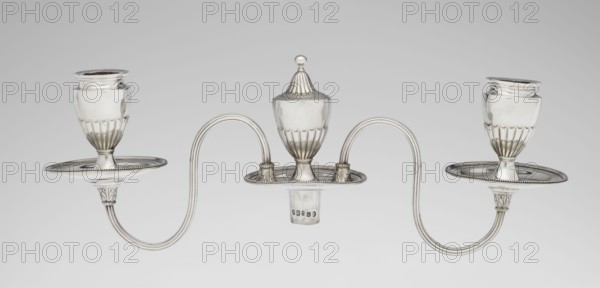 Two-candle candelabrum. Maker: Charles Hougham fl. 1769–1793. Style: Classicism, George III / Georgian. Description: Two-armed candelabrum on an oval foot bordered with pearling and decorated with a knobbed rosette. Conical, fluted stem topped with a vase-shaped socket, containing a setting with two S-shaped arms terminating in saucers and vase-shaped sockets with drip-pans; center between the arms is a vase topped with a ball. Sleeves decorated halfway with vertical flutes, smooth above. Date: 1787/1788. Place: London, England. Function: Lighting. Materials/technique: goldsmith work. Dimensions: height 38 cm, width 39 cm, depth 12 cm; weight 1060 g., A symmetrical metallic object with a shiny silver finish consisting of a central lidded vessel with a rounded finial on top flanked by two matching goblet-like cups, all set on shallow oval trays with beaded rims; the cups and central vessel have fluted lower sections and sit on short stems with decorated collars, and two sinuous double-a...