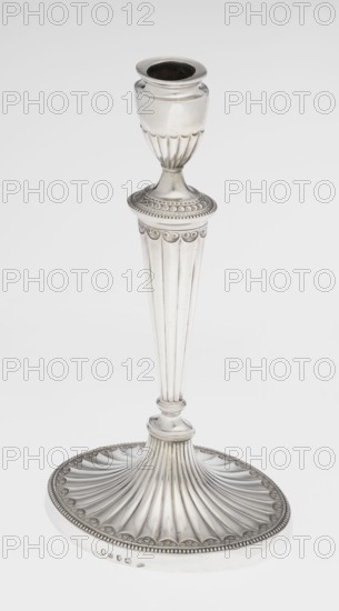 Two-candle candelabrum, maker: Charles Hougham fl. 1769–1793. Classicism/Georgian, George III style. Silver; fluted, pearling and rosette ornament. Two-armed candelabrum on an oval foot bordered with pearling and decorated with a knobbed rosette. Conical fluted stem topped with a vase-shaped socket containing a setting with two S-shaped arms terminating in saucers and vase-shaped sockets; central vase topped with a ball; sleeves decorated halfway with vertical flutes, smooth above. Dated 1787/1788. England, London. Function: lighting. Height 38 cm; width 39 cm; depth 12 cm; weight 1060 g., A tall metallic candlestick with a reflective silver surface standing on an ornate round base; the base has a fluted, fan-like pattern radiating outward and a beaded rim with a repeating scalloped motif, transitioning into a slender, vertically grooved stem that tapers upward to a decorative collar with circular embossed details, leading to a fluted cup-shaped holder at the top. The visible colors ar...