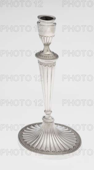 Two-candle candelabrum by Charles Hougham fl. 1769–1793. Classicism; George III/Georgian style. Two-armed candelabrum on an oval foot bordered with pearling and decorated with a knobbed rosette. Conical, fluted stem topped with a vase-shaped socket containing a setting with two S-shaped arms terminating in saucers and vase-shaped sockets with profiled fittings; between the arms is a vase topped with a ball. Sleeves decorated halfway with vertical flutes, smooth above. Materials: presumed silver; function: lighting. Date: 1787/1788. Place: London, England. Height 38 cm; width 39 cm; depth 12 cm; weight 1060 g., A tall metallic candlestick stands against a plain light background, showing a silvery surface with polished highlights and subtle darker reflections; the base is wide and round with fluted, fan-like ridges and a beaded decorative edge, the stem tapers upward with vertical fluting and a smooth rounded knop, and the top narrows to a cup-shaped holder with scalloped detailing benea...