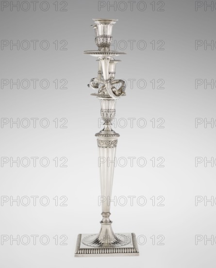 Classicist Two-Armed Candelabra, Schofield, John fl. 1776–1796 goldsmith, classicism style, godron ornaments, monograms, faceting, leaves ornament, Georgian, George III, Classicist two-armed candelabra. On a square base surrounded by godron ornament, passing into a circular foot decorated with a surrounding rosette; faceted stem enclosed at the top with a knurled floral tendril, terminating in a vase-shaped socket decorated with acanthus leaves and facets, edged with godrons, in which is set a setting in the form of two faceted, rotating arms ending with plate-shaped profiled dishes and vase-shaped candle holders, separated by a decorative vase with a lid. Engraved on several elements with a tied monogram PHB; dated 1786/1787, London, England, Great Britain. Function: lighting. Dimensions: height 47.5 cm, width 38 cm, depth 13 cm; weight 850 g., A tall metallic candlestick stands against a plain light gray background, showing a reflective silver surface with polished highlights and sub...