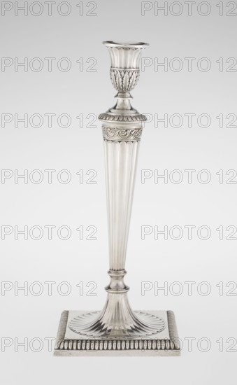 Classicist Two-Armed Candelabra, Schofield, John fl. 1776–1796 goldsmith; classicism, Georgian/George III style. Classicist two-armed candelabra on a square base surrounded by godron ornament, passing into a circular foot decorated with a surrounding rosette; faceted stem enclosed at the top with a knurled floral tendril, terminating in a vase-shaped socket decorated with acanthus leaves and facets, edged with godrons, in which is set a setting in the form of two faceted, rotating arms ending with plate-shaped profils and vase-shaped candle holders, separated by a decorative vase with a lid. Engraved on several elements with a tied monogram PHB, dated 1786/1787, London, England, Great Britain. Function: lighting. Dimensions: height 47.5 cm, width 38 cm, depth 13 cm; weight 850 g., A tall, slender metal candlestick with a fluted shaft and ornate detailing; the piece is predominantly silver in color with polished reflective surfaces. At the top there is a cup-like holder with vertical fl...