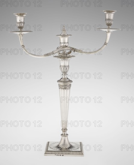 Classicist Two-Armed Candelabra, Schofield, John fl. 1776–1796 goldsmith, classicism, godron ornaments, monograms, faceting, leaves ornament, Georgian, George III. Classicist two-armed candelabra on a square base surrounded by godron ornament, passing into a circular foot decorated with a surrounding rosette; faceted stem enclosed at the top with a knurled floral tendril, terminating in a vase-shaped socket decorated with acanthus leaves and facets, edged with godrons, in which is set a setting in the form of two faceted, rotating arms ending with plate-shaped profites and vase-shaped candle holders, separated by a decorative vase with a lid. Engraved on several elements with a tied monogram PHB, 1786/1787, London, England, Great Britain. Function: lighting. Height 47.5 cm, width 38 cm, depth 13 cm, weight 850 g., A silver-colored metal two-armed candelabrum with a tall, fluted central stem standing on a square base; the base has a ridged border and a fluted, fan-like detail where it m...