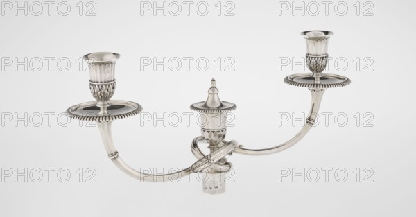 Classicist Two-Armed Candelabra, Schofield, John fl. 1776–1796 goldsmith; classicism, Georgian / George III style. Classicist two-armed candelabra on a square base surrounded by godron ornament, passing into a circular foot decorated with a surrounding rosette; faceted stem enclosed at the top with a knurled floral tendril, terminating in a vase-shaped socket decorated with acanthus leaves and facets, edged with godrons, in which is set a setting in the form of two faceted, rotating arms ending with plate-shaped profiled and vase-shaped candle holders, separated by a decorative vase with a lid. Engraved on several elements with a tied monogram PHB, dated 1786/1787, London, England, Great Britain. Function: lighting. Dimensions: height 47.5 cm, width 38 cm, depth 13 cm; weight 850 g., A silver three-armed candelabrum is shown against a pale gray background, with a central upright decorative finial and two curved arms extending outward, each terminating in a cup-shaped candle holder set ...