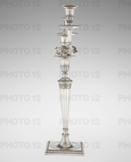 Classicist Two-Armed Candelabra, Schofield, John fl. 1776–1796 goldsmith; classicism, Georgian/George III style. Classicist two-armed candelabra on a square base surrounded by godron ornament, passing into a circular foot decorated with a surrounding rosette; faceted stem enclosed at the top with a knurled floral tendril, terminating in a vase-shaped socket decorated with acanthus leaves and facets, edged with godrons, in which is set a setting in the form of two faceted, rotating arms ending with plate-shaped profils and vase-shaped candle holders, separated by a decorative vase with a lid. Engraved on several elements with a tied monogram PHB, dated 1786/1787, London, England, Great Britain. Function: lighting. Dimensions: height 47.5 cm, width 38 cm, depth 13 cm; weight 850 g., A tall ornate metal candlestick stands against a neutral gray background; it has a square base with fluted and beaded edges, a tapered fluted column rising to an intricately molded central section with acanth...