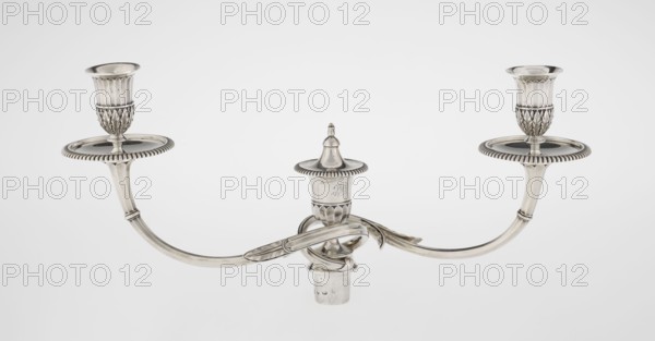 Classicist Two-Armed Candelabra, Schofield, John fl. 1776–1796 goldsmith, classicism, godron ornaments, monograms, faceting, leaves ornament, Georgian/George III style. Classicist two-armed candelabra on a square base surrounded by godron ornament, passing into a circular foot decorated with a surrounding rosette; faceted stem enclosed at the top with a knurled floral tendril, terminating in a vase-shaped socket decorated with acanthus leaves and facets, edged with godrons, containing two faceted, rotating arms ending with plate-shaped drip pans and vase-shaped candle holders, separated by a decorative vase with a lid. Engraved on several elements with a tied monogram PHB, dated 1786/1787, London, England, Great Britain. Function: lighting. Measurements: height 47.5 cm, width 38 cm, depth 13 cm; weight 850 g., A polished metallic three-armed candelabrum with a central finial and two matching candle cups at the ends of curved arms, all rendered in silver tones with reflective highlights...