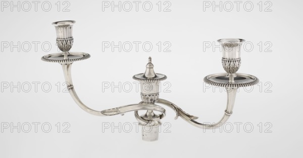 Classicist Two-Armed Candelabra, Schofield, John fl. 1776–1796 goldsmith, classicism, godron ornaments, monograms, faceting, leaves ornament, Georgian, George III. Classicist two-armed candelabra on a square base surrounded by godron ornament, passing into a circular foot decorated with a surrounding rosette; faceted stem enclosed at the top with a knurled floral tendril, terminating in a vase-shaped socket decorated with acanthus leaves and facets, edged with godrons, in which is set a setting in the form of two faceted, rotating arms ending with plate-shaped profils and vase-shaped candle holders, separated by a decorative vase with a lid. Engraved on several elements with a tied monogram PHB, 1786/1787, London, England, Great Britain. Function: lighting. Height 47.5 cm, width 38 cm, depth 13 cm, weight 850 g., A metallic three-arm candle holder with a central finial and two outward-curving arms ending in raised circular drip plates and cup-shaped holders; the metal surface is polish...