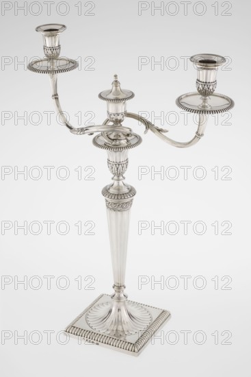 Classicist Two-Armed Candelabra, Schofield, John fl. 1776–1796 goldsmith; classicism, Georgian/George III styles. Classicist two-armed candelabra on a square base surrounded by godron ornament, passing into a circular foot decorated with a surrounding rosette; faceted stem enclosed at the top with a knurled floral tendril, terminating in a vase-shaped socket decorated with acanthus leaves and facets, edged with godrons, in which is set a setting in the form of two faceted, rotating arms ending with plate-shaped profilers and vase-shaped candle holders, separated by a decorative vase with a lid. Engraved on several elements with a tied monogram PHB, dated 1786/1787, London, England. Lighting; height 47.5 cm, width 38 cm, depth 13 cm, weight 850 g., A silver-colored three-armed candelabrum stands on a square base with ridged edging and a fluted, scalloped central platform; a tall, tapered fluted column rises from the base, topped by an ornate capital with ribbed and leaf-like patterns, s...