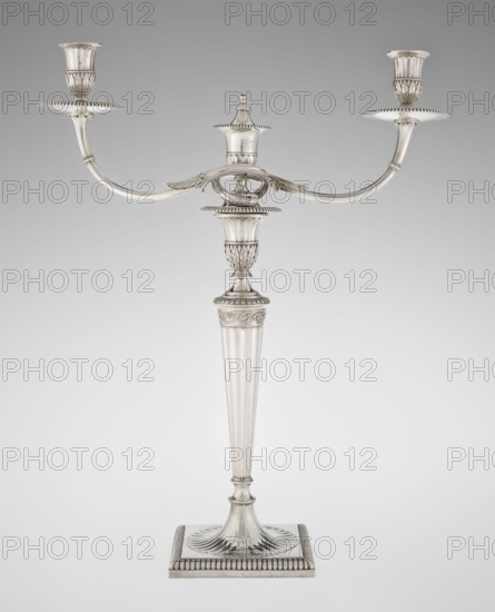 Classicist Two-Armed Candelabra, Schofield, John fl. 1776–1796 goldsmith, classicism style, Georgian / George III style. Classicist two-armed candelabra on a square base surrounded by godron ornament, passing into a circular foot decorated with a surrounding rosette; faceted stem enclosed at the top with a knurled floral tendril, terminating in a vase-shaped socket decorated with acanthus leaves and facets, edged with godrons, in which is set a setting in the form of two faceted, rotating arms ending with plate-shaped profils and vase-shaped candle holders, separated by a decorative vase with a lid. Engraved on several elements with a tied monogram PHB, dated 1786/1787, London, Great Britain. Function: lighting. Dimensions: height 47.5 cm, width 38 cm, depth 13 cm; weight 850 g., A tall metallic candelabrum with a reflective silver surface stands against a neutral gray background; it has a square base with a fluted, scalloped design rising to a tapered, fluted column topped by ornate a...