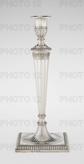 Classicist Two-Armed Candelabra, Schofield, John fl. 1776–1796 goldsmith, classicism, Georgian / George III style. Classicist two-armed candelabra on a square base with godron ornament, passing into a circular foot decorated with a surrounding rosette; faceted stem enclosed at the top with a knurled floral tendril, terminating in a vase-shaped socket decorated with acanthus leaves and facets, edged with godrons. Set in the socket are two faceted, rotating arms ending with plate-shaped profits and vase-shaped candle holders, separated by a decorative vase with a lid. Engraved on several elements with a tied monogram PHB; dated 1786/1787. Place: London, England. Function: lighting. Dimensions: height 47.5 cm, width 38 cm, depth 13 cm; weight 850 g., A single tall metallic candlestick stands against a plain light background, rendered in silver tones with reflective highlights and subtle darker gray shadows. The candlestick has a flared cup at the top with vertical fluting, a decorative te...