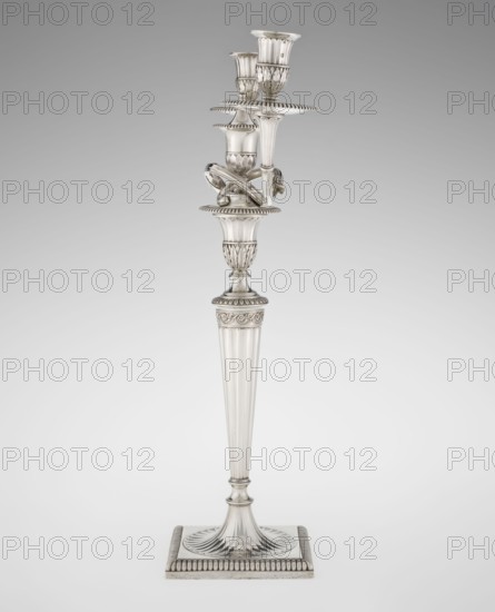 Classicist two-armed candelabra by John Schofield fl. 1776–1796, goldsmith; classicism/Georgian/George III style. On a square base surrounded by godron ornament, passing into a circular foot decorated with a surrounding rosette; faceted stem enclosed at the top with a knurled floral tendril, terminating in a vase-shaped socket decorated with acanthus leaves and facets, edged with godrons, in which is set a setting in the form of two faceted, rotating arms ending with plate-shaped profils and vase-shaped candle holders, separated by a decorative vase with a lid. Engraved on several elements with a tied monogram PHB, dated 1786/1787, made in London, England. Lighting object; height 47.5 cm, width 38 cm, depth 13 cm, weight 850 g., A tall metallic candlestick with a reflective silver surface stands on a square base with a ridged border and a fluted circular platform, the stem above it tapering upward with vertical fluting and decorative bands, leading to a carved collar with a leaf-like m...