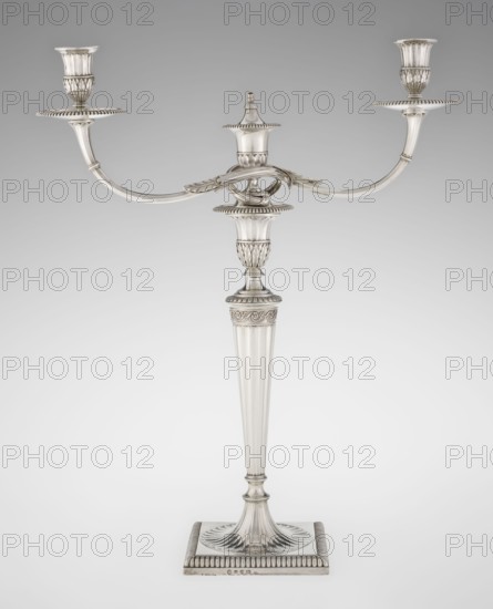 Classicist Two-Armed Candelabra, by John Schofield fl. 1776–1796, goldsmith; Classicism/Georgian/George III style. Classicist two-armed candelabra on a square base surrounded by godron ornament, passing into a circular foot decorated with a surrounding rosette; faceted stem enclosed at the top with a knurled floral tendril, terminating in a vase-shaped socket decorated with acanthus leaves and facets, edged with godrons, in which is set a setting in the form of two faceted, rotating arms ending with plate-shaped profils and vase-shaped candle holders, separated by a decorative vase with a lid. Decorated with godron ornaments, faceting, leaves ornament and engraved on several elements with a tied monogram PHB, dated 1786/1787, London, Great Britain. Function: lighting. Dimensions: height 47.5 cm, width 38 cm, depth 13 cm; weight 850 g., A silver candelabrum stands against a neutral light gray background, featuring a tall fluted central stem rising from a square base with ridged edges an...