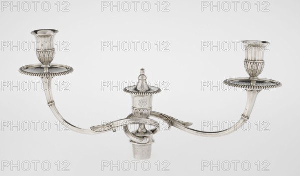 Classicist Two-Armed Candelabra, Schofield, John fl. 1776–1796, maker goldsmith; classicism, Georgian/George III style. Classicist two-armed candelabra on a square base surrounded by godron ornament, passing into a circular foot decorated with a surrounding rosette; faceted stem enclosed at the top with a knurled floral tendril, terminating in a vase-shaped socket decorated with acanthus leaves and facets, edged with godrons, in which is set a setting in the form of two faceted, rotating arms ending with plate-shaped profils and vase-shaped candle holders, separated by a decorative vase with a lid. Engraved on several elements with a tied monogram PHB, dated 1786/1787, London, England. Function: lighting. Dimensions: height 47.5 cm, width 38 cm, depth 13 cm; weight 850 g., A silver three-armed candelabrum with a central fluted finial and two curved arms extending outward, each arm ending in a decorative holder with a fluted cup and a round drip plate with a beaded edge; the metal surfa...