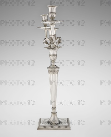 Classicist Two-Armed Candelabra, Schofield, John fl. 1776–1796 goldsmith, classicism style, godron ornaments, monograms, faceting, leaves ornament, Georgian, George III. Classicist two-armed candelabra on a square base surrounded by godron ornament, passing into a circular foot decorated with a surrounding rosette; faceted stem enclosed at the top with a knurled floral tendril, terminating in a vase-shaped socket decorated with acanthus leaves and facets, edged with godrons, in which is set a setting in the form of two faceted, rotating arms ending with plate-shaped profits and vase-shaped candle holders, separated by a decorative vase with a lid. Engraved on several elements with a tied monogram PHB, 1786/1787, England, London, Great Britain; function: lighting. Height 47.5 cm, width 38 cm, depth 13 cm, weight 850 g., A tall ornate metal candlestick stands against a plain background, with a fluted tapered shaft rising from a square base that has a ridged border and a circular fluted p...