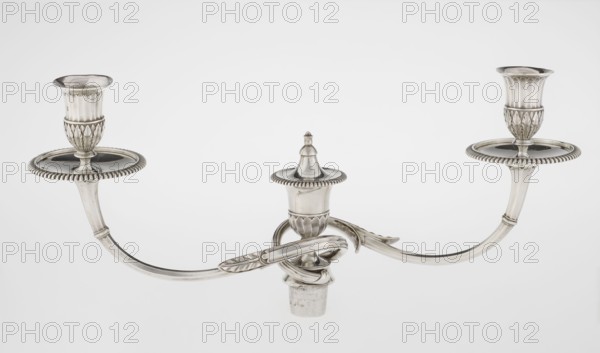 Classicist Two-Armed Candelabra, Schofield, John fl. 1776–1796 goldsmith; classicism, Georgian/George III style. Classicist two-armed candelabra on a square base surrounded by godron ornament, passing into a circular foot decorated with a surrounding rosette; faceted stem enclosed at the top with a knurled floral tendril, terminating in a vase-shaped socket decorated with acanthus leaves and facets, edged with godrons, in which is set a setting in the form of two faceted, rotating arms ending with plate-shaped profils and vase-shaped candle holders, separated by a decorative vase with a lid. Engraved on several elements with a tied monogram PHB, dated 1786/1787, London, Great Britain. Function: lighting. Dimensions: height 47.5 cm, width 38 cm, depth 13 cm; weight 850 g., A silver-colored metal two-armed candle holder with a central decorative finial and a curved, intertwined arm design, showing a few fluted and leaf-like ornamental details and round drip trays beneath each candle cup,...