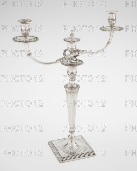 Classicist two-armed candelabra by John Schofield fl. 1776–1796, goldsmith; classicism/Georgian/George III style. On a square base surrounded by godron ornament, passing into a circular foot decorated with a surrounding rosette; faceted stem enclosed at the top with a knurled floral tendril, terminating in a vase-shaped socket decorated with acanthus leaves and facets, edged with godrons, containing two faceted, rotating arms ending with plate-shaped profils and vase-shaped candle holders, separated by a decorative vase with a lid. Engraved on several elements with a tied monogram PHB, dated 1786/1787. London, England. Lighting. Height 47.5 cm, width 38 cm, depth 13 cm, weight 850 g., A silver-colored metal candelabrum with a central fluted stem rising from a square base that has a ridged edge and a ridged, fan-like circular detail where the stem meets the base; the stem features decorative fluting and a few carved ornamental bands, leading to a central platform with a small domed fini...