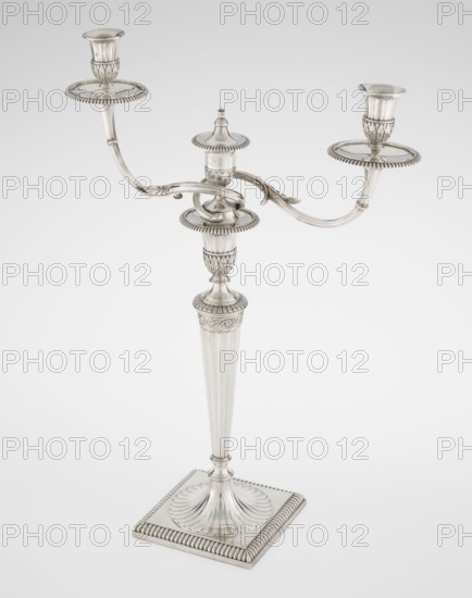 Classicist two-armed candelabra by John Schofield fl. 1776–1796, goldsmith. Classicism/Georgian George III style. On a square base surrounded by godron ornament, passing into a circular foot decorated with a surrounding rosette; faceted stem enclosed at the top with a knurled floral tendril, terminating in a vase-shaped socket decorated with acanthus leaves and facets, edged with godrons, containing two faceted, rotating arms ending with plate-shaped profiled and vase-shaped candle holders, separated by a decorative vase with a lid. Engraved on several elements with a tied monogram PHB, dated 1786/1787, London, England. Material: silver lighting. Dimensions: height 47.5 cm, width 38 cm, depth 13 cm; weight 850 g., A silver-colored three-armed candelabrum standing on a square base with fluted and beaded decorative edges, a tapered fluted central column rising to an ornate central socket, and two curved arms extending outward to hold cup-shaped candle sockets atop round drip trays, all s...