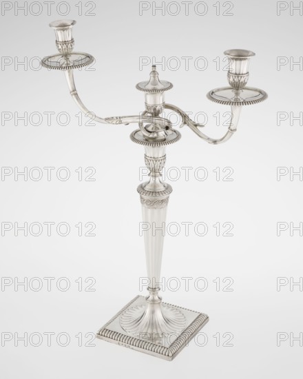 Classicist Two-Armed Candelabra, Schofield, John fl. 1776–1796 goldsmith, classicism style, Georgian / George III style. Classicist two-armed candelabra on a square base surrounded by godron ornament, passing into a circular foot decorated with a surrounding rosette; faceted stem enclosed at the top with a knurled floral tendril, terminating in a vase-shaped socket decorated with acanthus leaves and facets, edged with godrons, in which is set a setting in the form of two faceted, rotating arms ending with plate-shaped profils and vase-shaped candle holders, separated by a decorative vase with a lid. Engraved on several elements with a tied monogram PHB, dated 1786/1787, London, England. Function: lighting. Height 47.5 cm, width 38 cm, depth 13 cm, weight 850 g., A silver three-armed candelabrum stands against a plain light gray background, showing a central fluted column rising from a square base with a carved shell-like motif and a beaded edge; the column and base are metallic silver ...