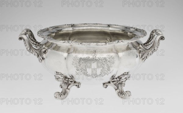 Punch vase, maker: Paul Storr 1771–1844; firm: Storr & Mortimer London; goldsmiths, fl. 1822–1838. English coat of arms engraved on the belly in a decorative cartouche. Materials/ornament: acanthus, shells, volutes; burling; burled and ribbed spherical, flattened eight-part form; supported on four volute leafy feet; two handles decorated with acanthus leaves and stylized shells. Upper edge turned up and decorated with relief ornament of leaves and shells. Function: punch vessel. Date: 1831/1832. Place of origin: London, England, Great Britain. Vessel. Height 15.5 cm; width 36.2 cm; depth 27.8 cm; weight 2490 g., A polished metal bowl with ornate scrolling handles and feet sits against a neutral background, showing a reflective surface in shades of silver and gray with darker reflections; the bowl has a scalloped rim and a smooth rounded body with decorative engraved flourishes and a central embellished cartouche, and a few intricate leaf and scroll motifs appear on the handles and feet...