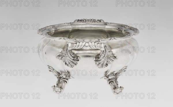Punch vase, by Paul Storr 1771–1844 and Storr & Mortimer London; goldsmiths, fl. 1822–1838. Spherical, flattened, eight-part, burled and ribbed punch vase with burling; supported on four volute leafy feet. Two handles decorated with acanthus leaves on the sides and stylized shells. Upper edge turned up and decorated with relief ornament of leaves and shells. Engraved coat of arms on the belly in a decorative cartouche. Materials/ornament: acanthus, shells, volutes. Function: punch vessel. Date: 1831/1832. Origin: London, England, Great Britain. Dimensions: height 15.5 cm; width 36.2 cm; depth 27.8 cm; weight 2490 g., A polished silver bowl with ornate curved edges and decorative handles sits on a plain light background; the bowl has a rounded, bulbous body with intricate leaf-like embellishments near the handles and on the scrolling feet, and the interior and exterior surfaces show reflective highlights and soft shadows in silver and gray tones.