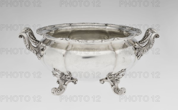 Punch vase by Paul Storr 1771–1844 and Storr & Mortimer London; goldsmiths, fl. 1822–1838. English, 1831/1832. Silver punch vessel, spherical flattened eight-part, burled and ribbed with burling and ribbing; supported on four volute leafy feet. Two handles decorated with acanthus leaves and stylized shells. Upper edge turned up and decorated with relief ornament of leaves and shells. Belly engraved with a coat of arms in a decorative cartouche. Materials/technique: silver; motifs include acanthus, shells, volutes. Dimensions: height 15.5 cm, width 36.2 cm, depth 27.8 cm; weight 2490 g., A shiny silver bowl with ornate scrolling handles and feet sits against a pale gray background, the bowl's rounded body reflecting light and showing smooth and slightly dimpled surfaces, the rim decorated with carved floral and leaf motifs, the handles and feet featuring elaborate acanthus-like scrollwork and textured detailing in the same silver tone, and the overall colors visible are silver and shade...